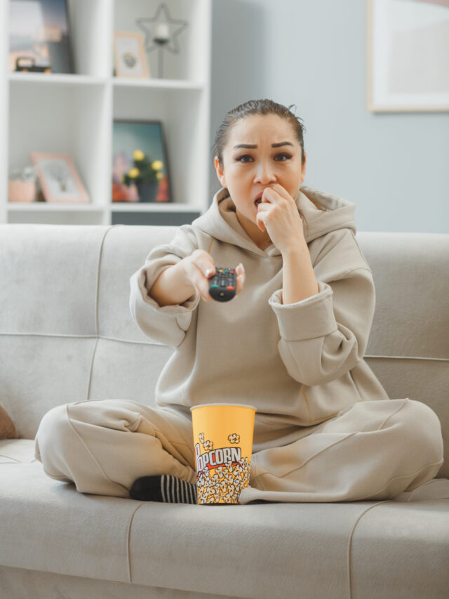 young beautiful woman in home clothes sitting on a couch at home interior with bucket of popcorn holding remote watching television being nervous and stressed biting nails