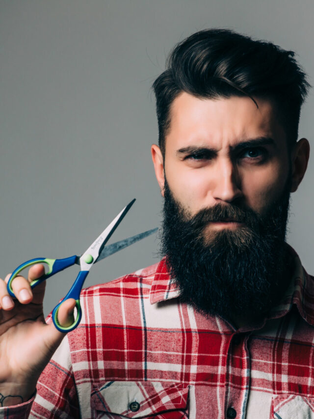 young handsome bearded man with long beard moustache and brunette hair holding hairdresser or barber scissors with emotional face in studio on grey background